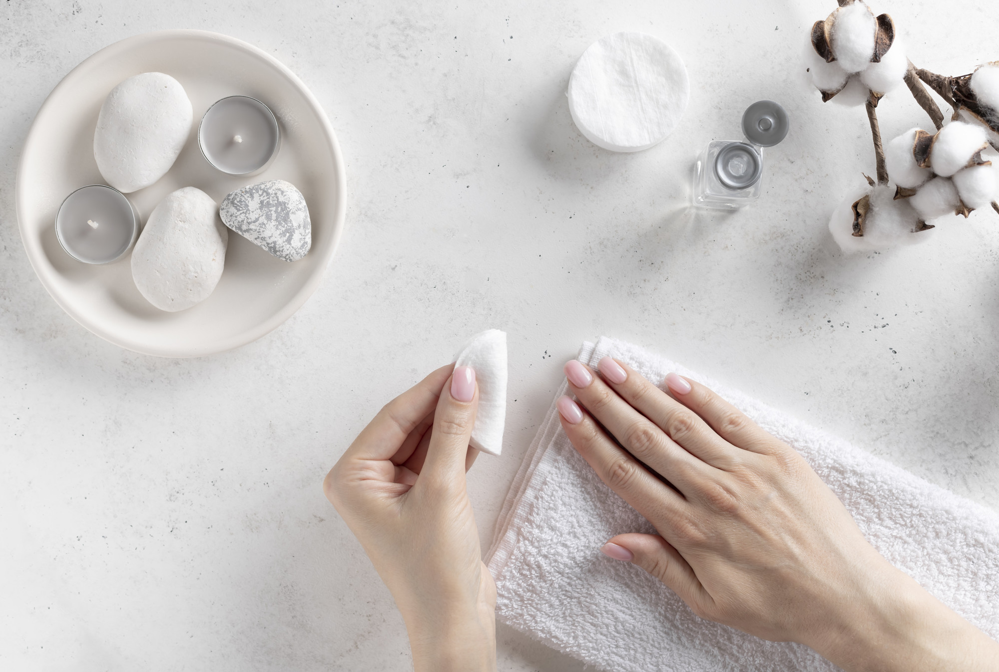 woman's hands holding a cotton pad and removing pink nail polish. hand care, manicure process. white concrete background, top view.